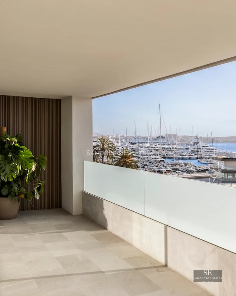 Spacious balcony with sea view. Glass railing, wooden floor, modern furniture, and lush vegetation.