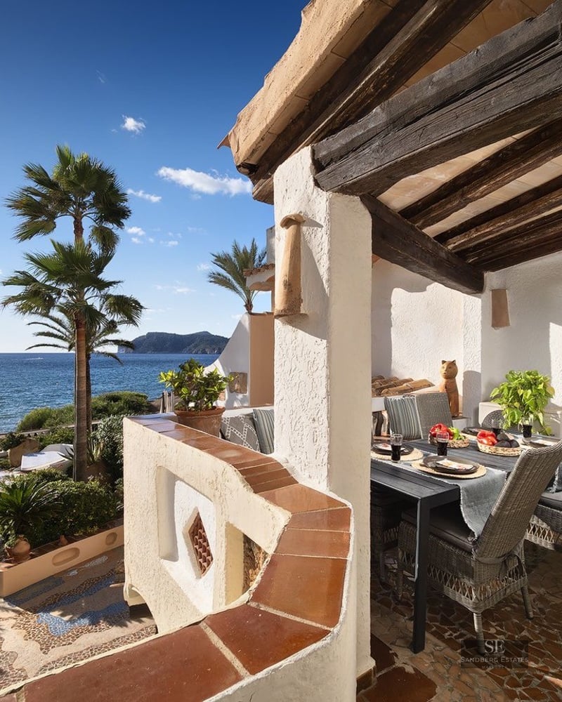 Covered terrace with rustic wood beams, dining table, wicker chairs, and a stunning view of the sea and palm trees.