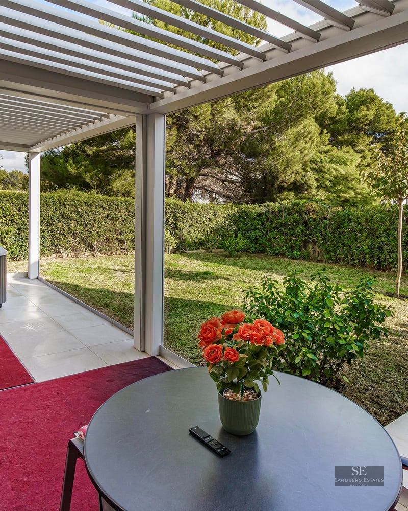 Outdoor terrace with a white pergola, grey hot tub, red rugs, and a view of a green garden with hedges.
