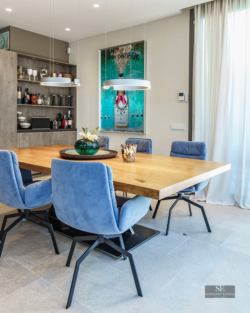 Salle à manger moderne avec table en bois, chaises bleues, cave à vin et grande baie vitrée ouvrant sur le jardin.
