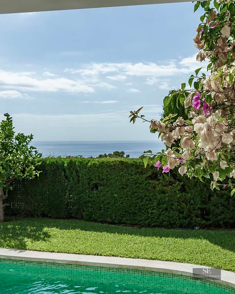 Turquoise swimming pool surrounded by green lawn and blooming flowers with panoramic sea views.