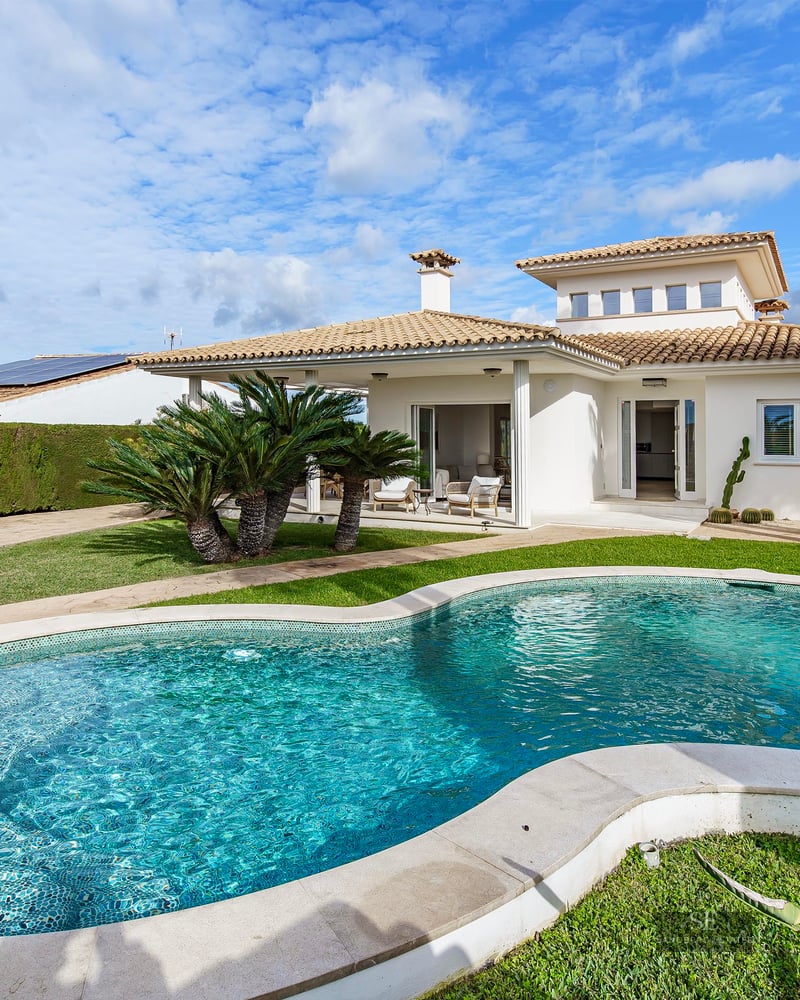 A turquoise kidney-shaped swimming pool surrounded by green grass and palm trees in front of a Mediterranean white villa.
