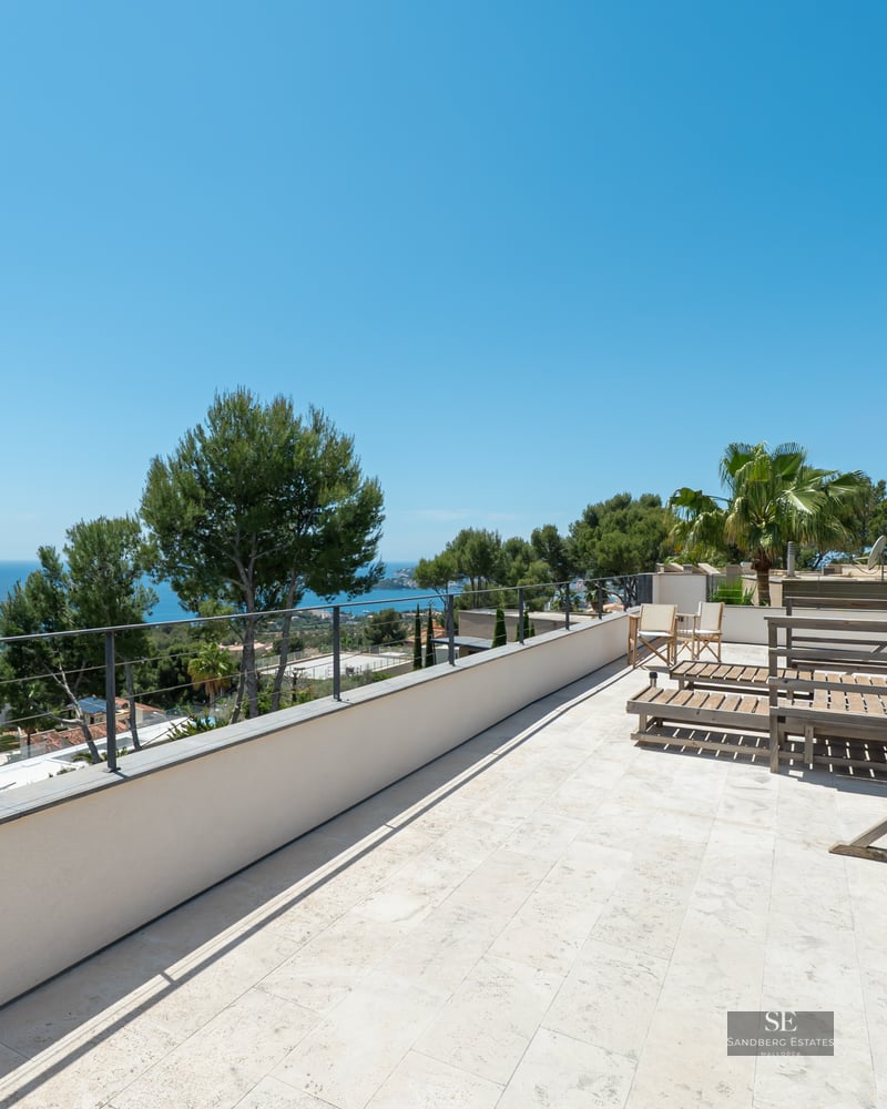 Large stone terrace with wooden loungers overlooking the Mediterranean Sea and lush trees.