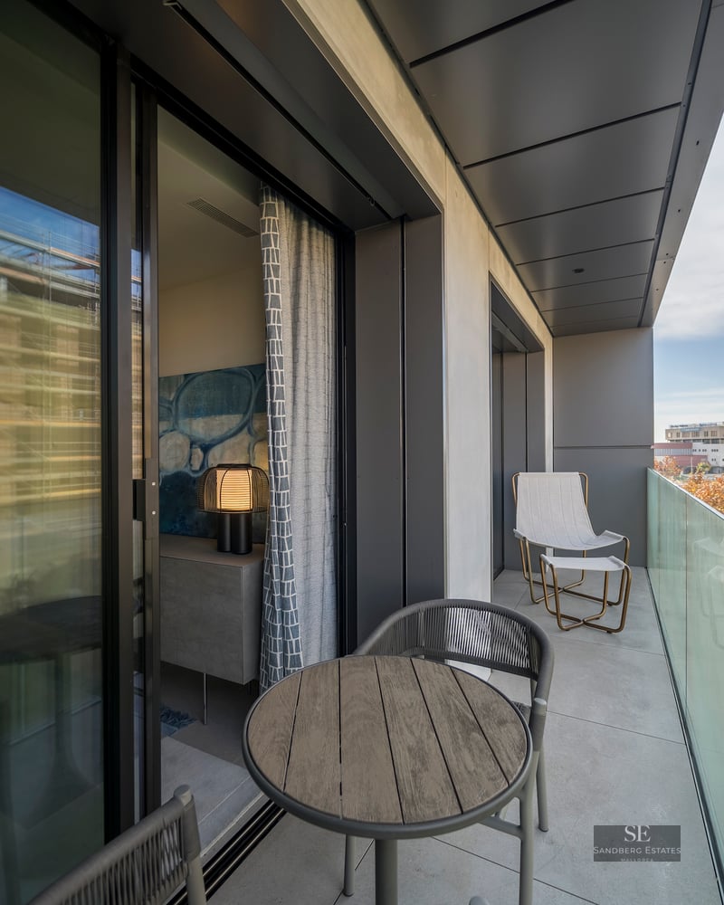A contemporary balcony with a glass railing, wooden table, and comfortable seating overlooking a green park area.
