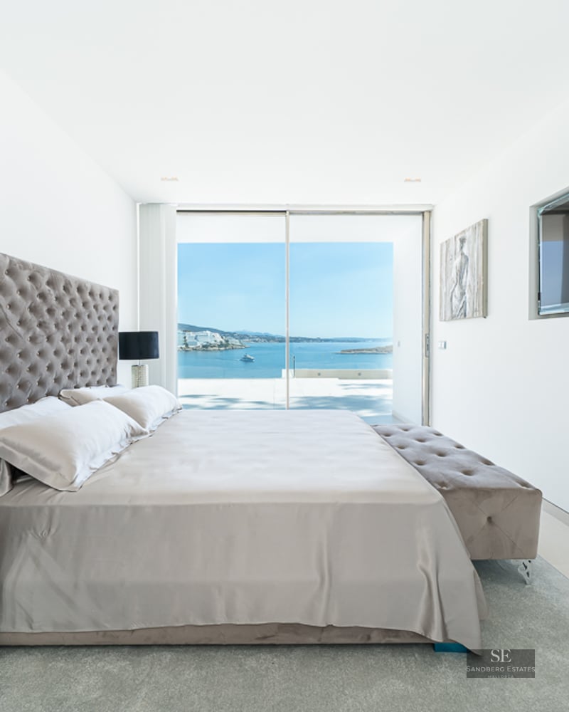 Modern master bedroom with tufted headboard, mirrored furniture, and large glass doors overlooking a blue ocean.