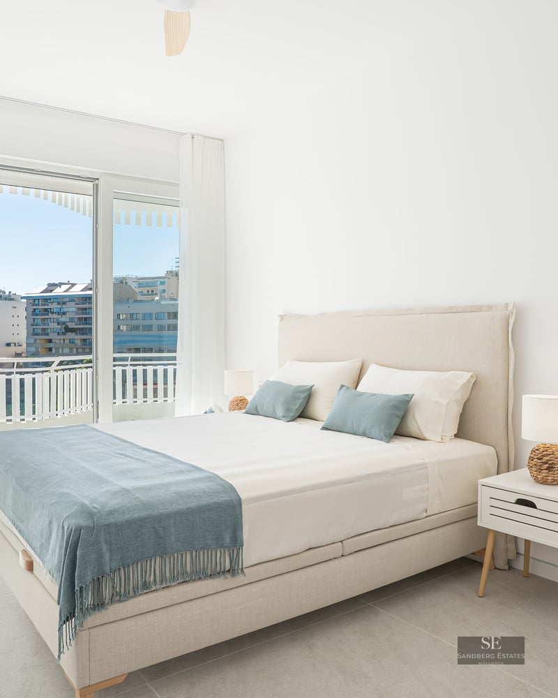 Bright bedroom with a double bed, bedside tables, and large glass doors opening to a balcony with a city view.