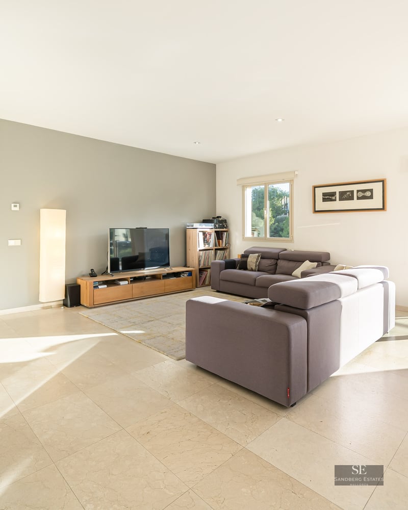Bright modern living room featuring grey sofas, a wooden dining table, and large windows overlooking a lush green forest.