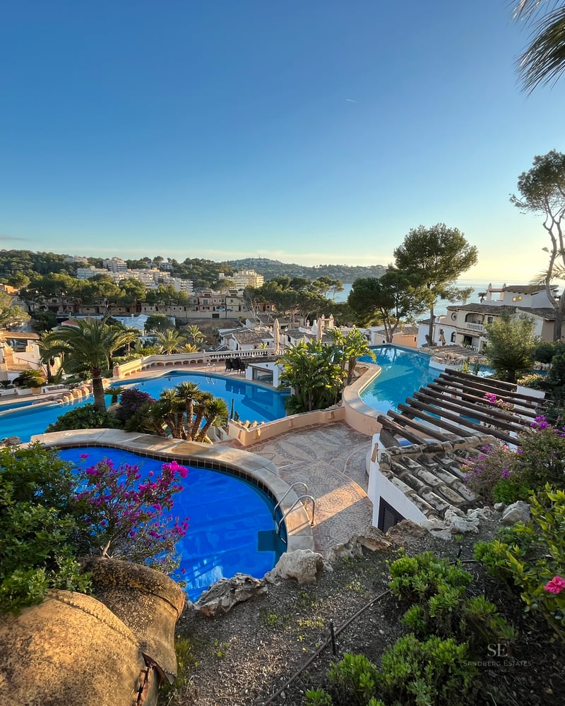 Multiple tiered blue swimming pools surrounded by lush tropical gardens and Mediterranean villas at sunset.