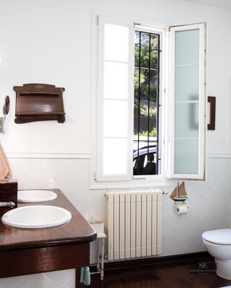 A bathroom featuring a double white sink vanity on dark wood, white tiled walls, a window, and a white toilet.