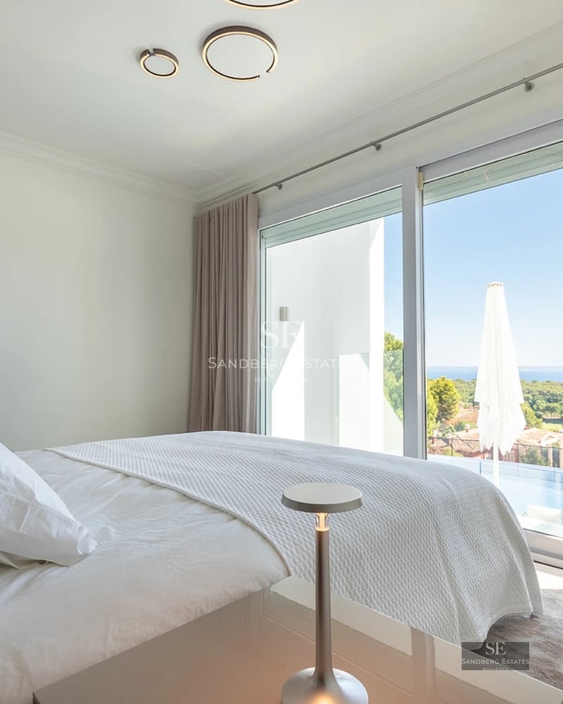 Bright master bedroom with a white bed and floor-to-ceiling glass doors overlooking a pool and the sea.