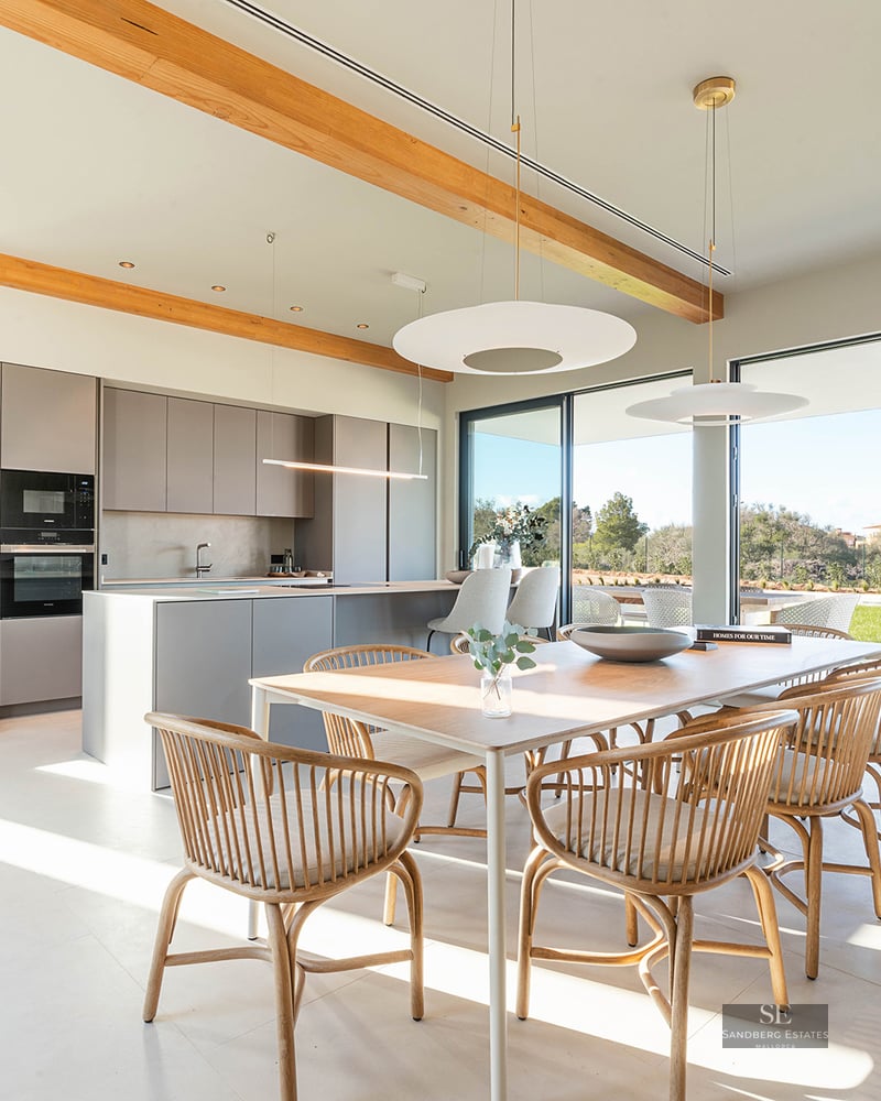 Bright dining room with wooden table and chairs next to a minimalist kitchen with large glass doors to garden.