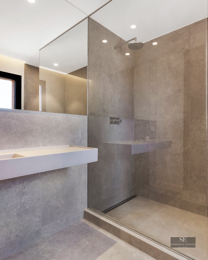 Minimalist bathroom with gray stone tiles, glass walk-in shower, wall-mounted sink, and large mirror.