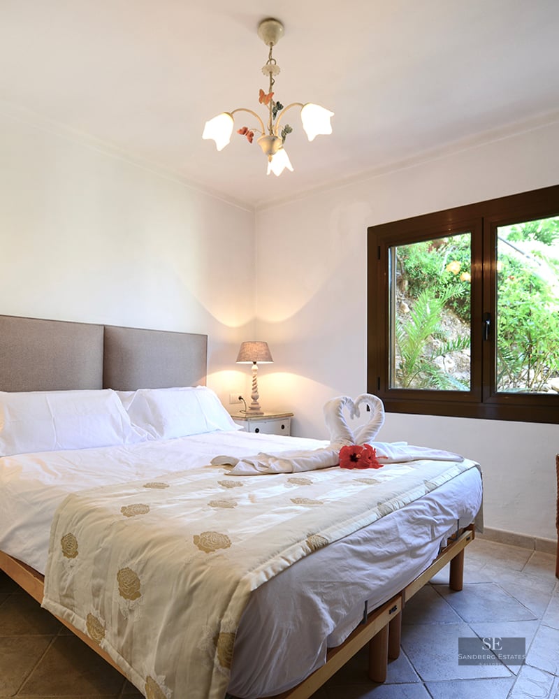 Bright bedroom with double bed, rustic nightstands, wicker chair, and window viewing lush outdoor greenery.