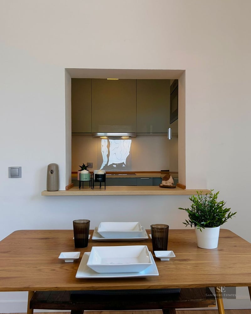 Wooden dining table set for two in front of a wall opening looking into a modern kitchen.