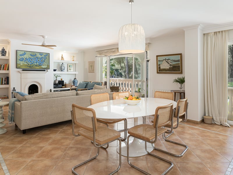 Open-plan living and dining room with terracotta tiles, white oval table, cane chairs, and views of pine trees.