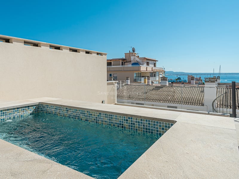 A rooftop plunge pool with beige stone coping and blue mosaic tiles, overlooking traditional rooftops and the sea.