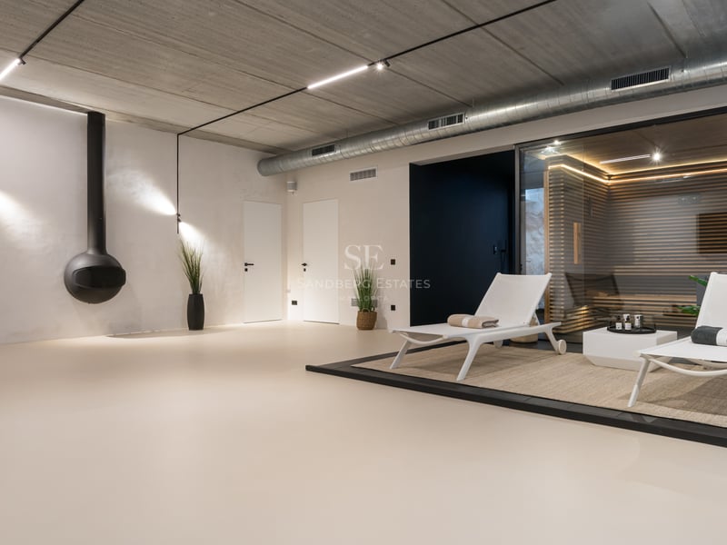 Minimalistiskt inomhus-spa med inglasad bastu, vit solstol och industriell inredning.