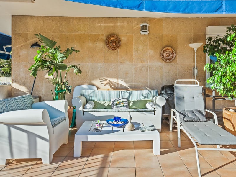 Sonnige überdachte Terrasse mit weißen Loungemöbeln, einer strukturierten Steinwand und üppigen Topfpflanzen.