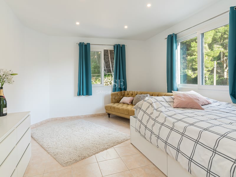 Chambre lumineuse avec lit, canapé en velours moutarde, rideaux bleu canard et vue sur la forêt par les fenêtres.