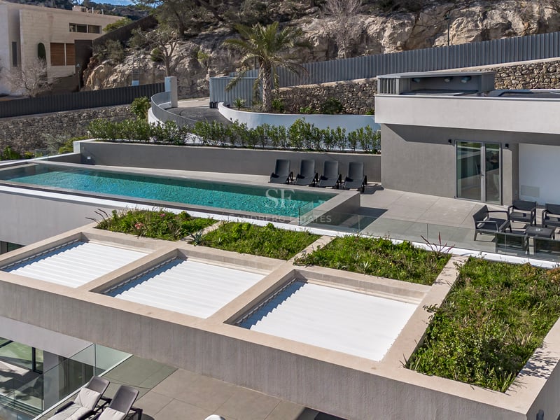 Vue aérienne d'une villa moderne grise avec piscine à débordement, chaises longues et toit végétalisé devant une colline.