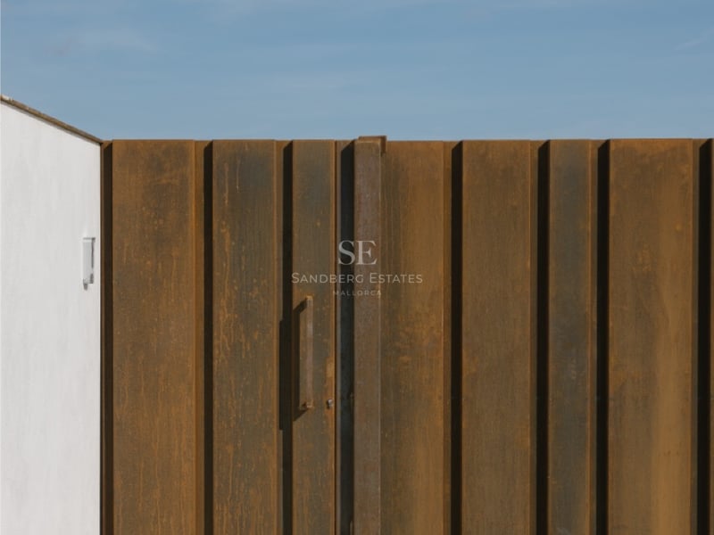 Vertikales Tor aus Cortenstahl mit minimalistischem Griff neben einer weißen Wand unter blauem Himmel.
