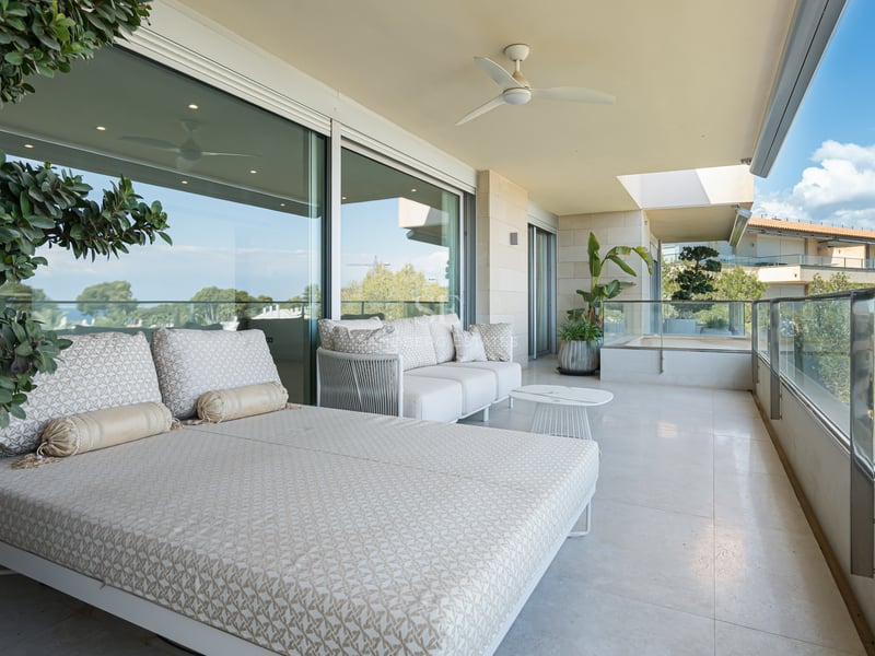 Large covered terrace with patterned daybed, glass railings, and views of lush greenery.