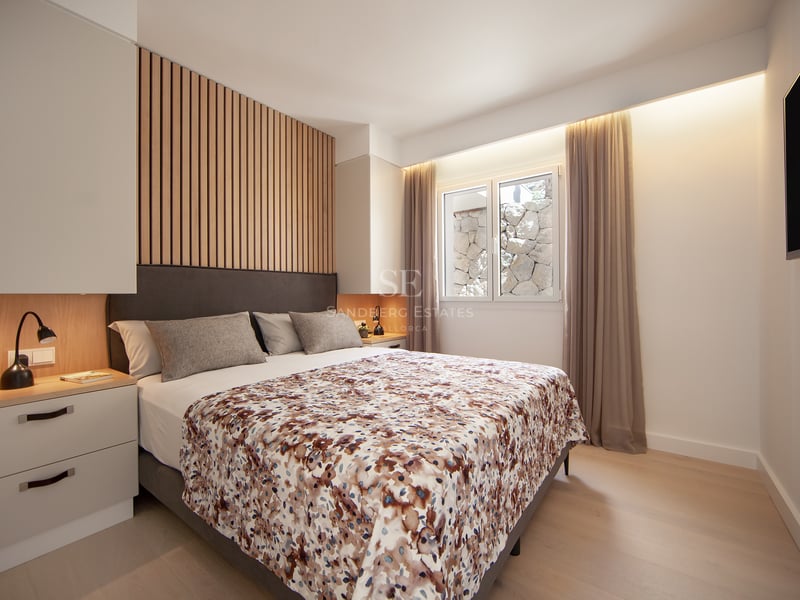 Chambre moderne avec lit gris, détail de mur en bois, éclairage chaleureux et fenêtre avec rideaux.