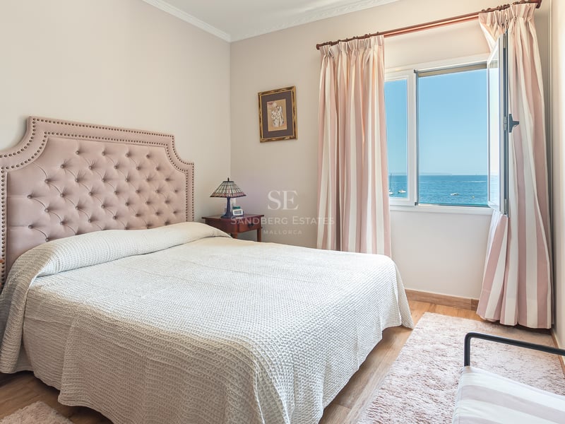 Ljust sovrum med rosa stoppad huvudgavel, trämöbler och fönster som öppnar upp mot det blå Medelhavet.