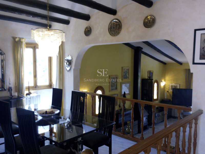 Matsal på övre plan med glasbord, mörka träbjälkar och utsikt över det nedre vardagsrummet.