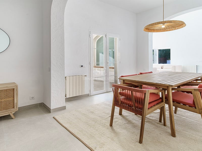 Ljus matsal med träbord, stolar med terrakottafärgade dynor, flätad taklampa och vita valv.