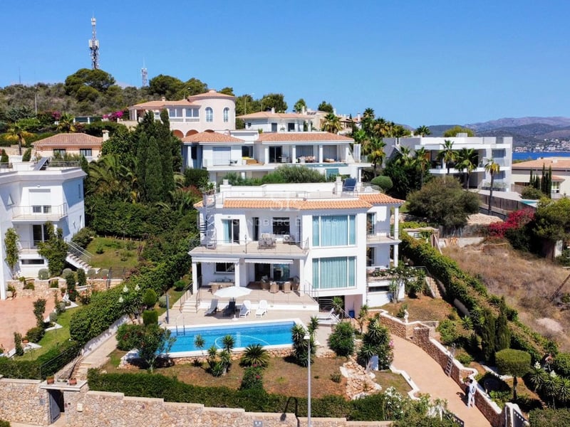 Vue aérienne d'une villa blanche à plusieurs niveaux avec une piscine turquoise, des murs en pierre et des jardins.