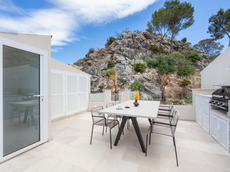 Terrasse blanche moderne avec table à manger, chaises et barbecue, avec une montagne rocheuse en arrière-plan.