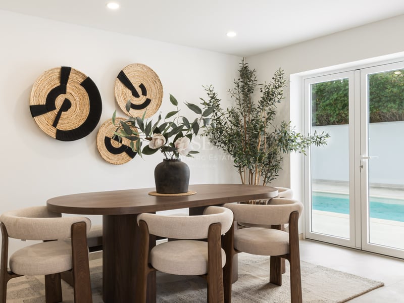 Table à manger ovale en bois avec chaises beiges devant des portes vitrées donnant sur une piscine.