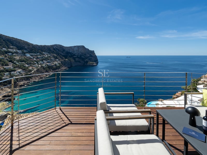 Elevated wooden terrace with lounge seating overlooking a Mediterranean bay and rugged coastline under a blue sky.