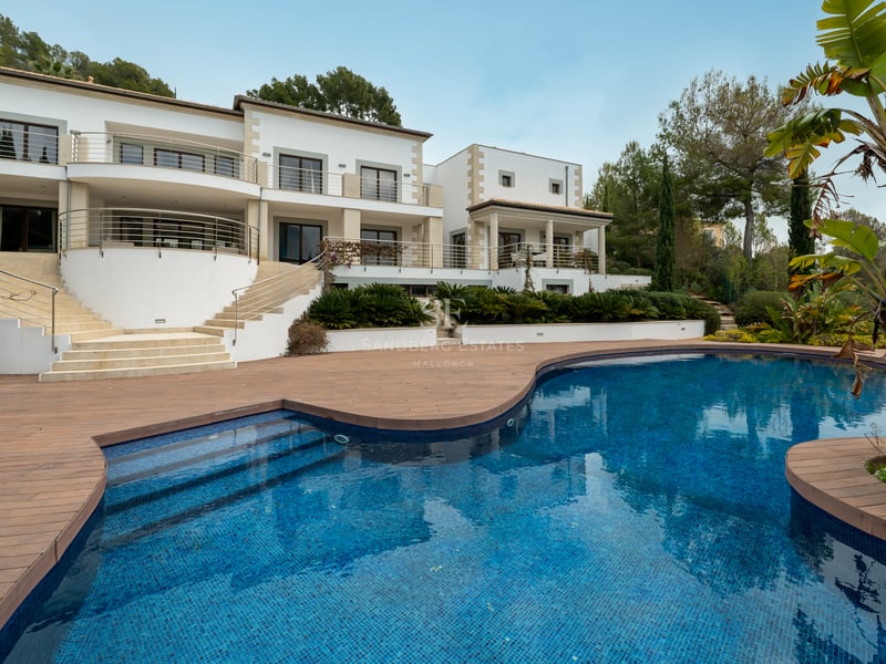 Vue extérieure d'une villa blanche de luxe avec une grande piscine courbe et terrasse en bois entourée de verdure.