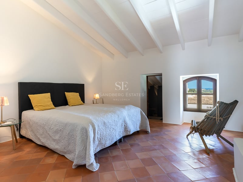 Chambre spacieuse avec poutres blanches, sol en terre cuite et fenêtre cintrée donnant sur la mer.