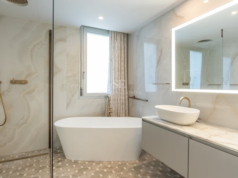 Luxury bathroom featuring marble walls, a freestanding white tub, brushed gold fixtures, and a large backlit mirror.