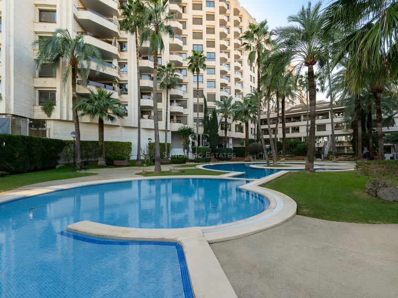 Slingrande blå pool omgiven av grönt gräs och höga palmer framför ett modernt lägenhetshus.