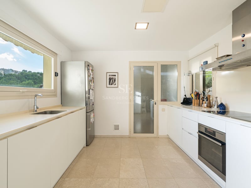 Ljust kök med vita skåp, rostfri kyl och ugn, samt ett stort fönster med utsikt över grönska.