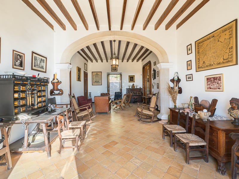 Vaste salon avec sols en terre cuite, poutres en bois apparentes, une grande arche en pierre et des meubles anciens.
