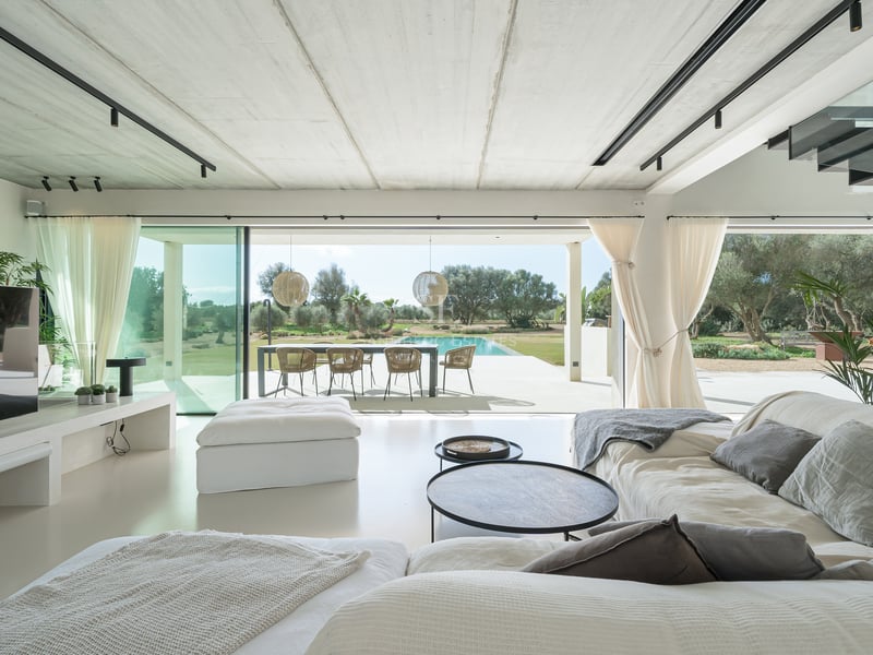 Ljust vardagsrum med minimalistisk inredning och stora glasdörrar mot en utomhuspool och matplats.