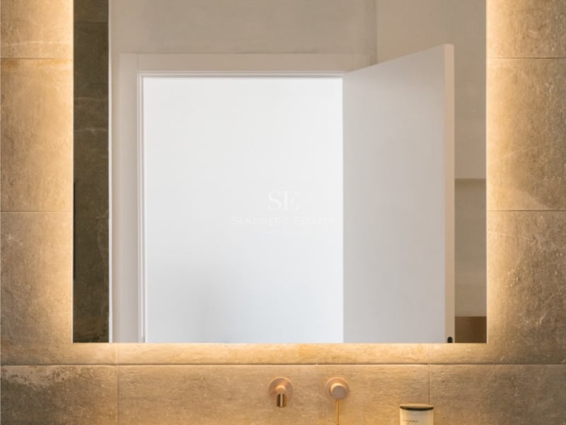 Nahaufnahme eines Waschtischs aus Naturstein mit integriertem Becken, Wandarmaturen aus Bronze und LED-Spiegel.