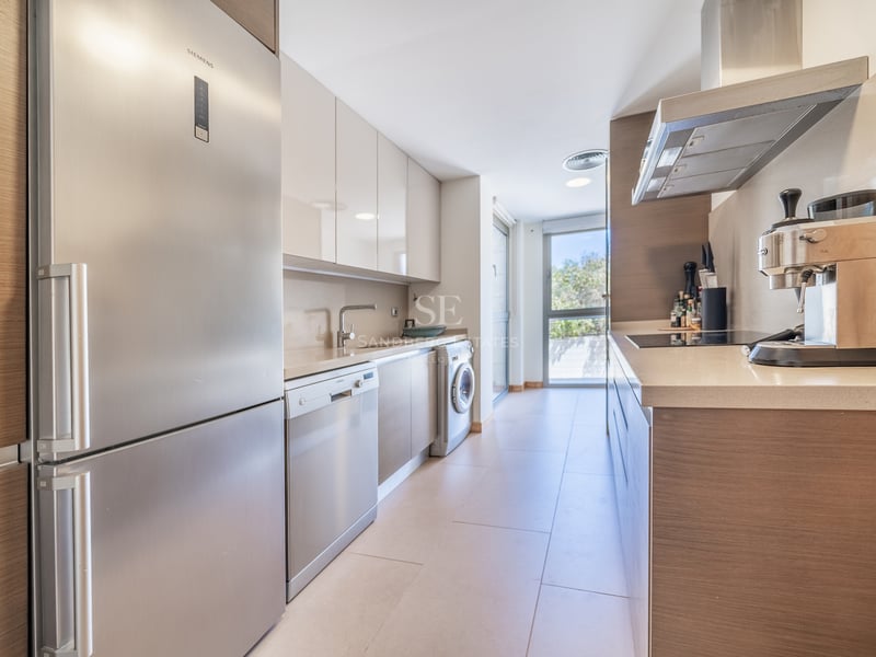 Cuisine en couloir avec placards en bois clair, appareils en inox et grande fenêtre au bout.