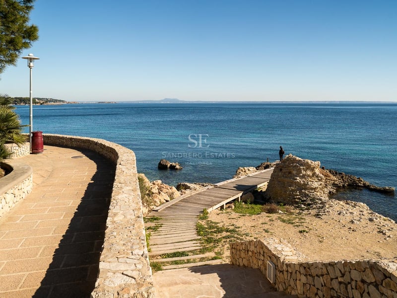 Küstenpromenade aus Naturstein und Holzsteg zum türkisfarbenen Mittelmeer unter blauem Himmel.