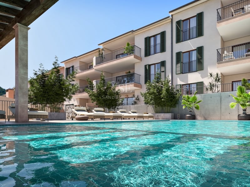 Large turquoise swimming pool with sun loungers in front of an apartment building with green shutters and balconies.