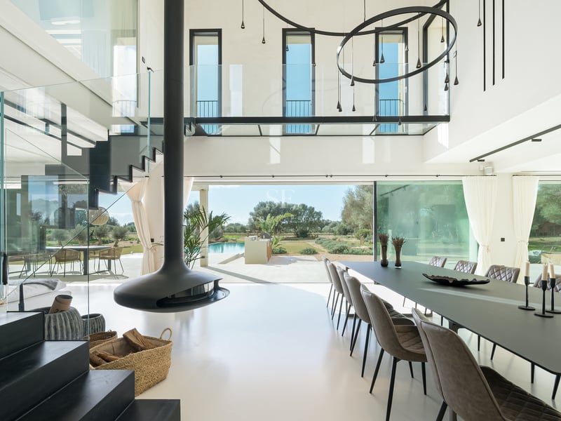 Ljus matsal med dubbel takhöjd, svart hängande öppen spis, glasräcken och utsikt över trädgård och pool.