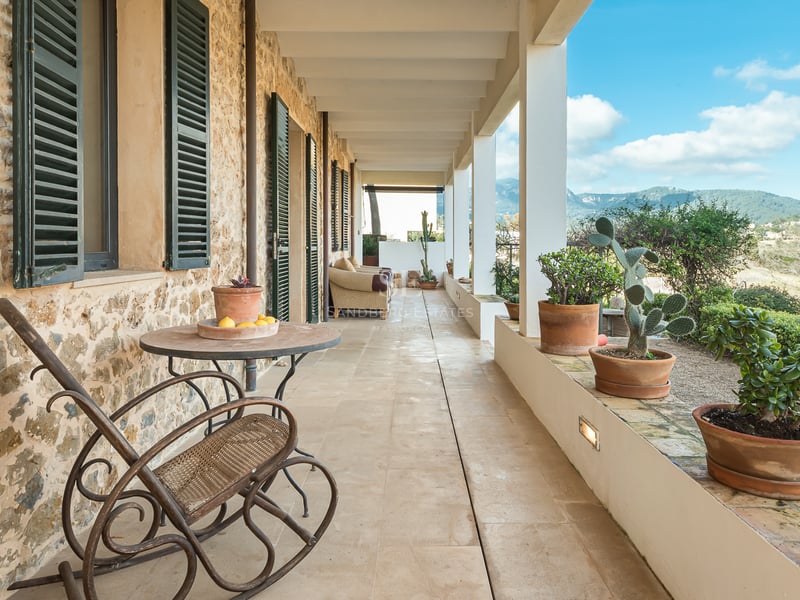 Täckt stenterrass med terrakottakrukor, gröna fönsterluckor och en gungstol med utsikt över bergen.