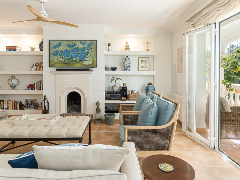 Bright living room with terracotta floors, white fireplace, built-in shelves, and glass doors opening to a lush terrace.