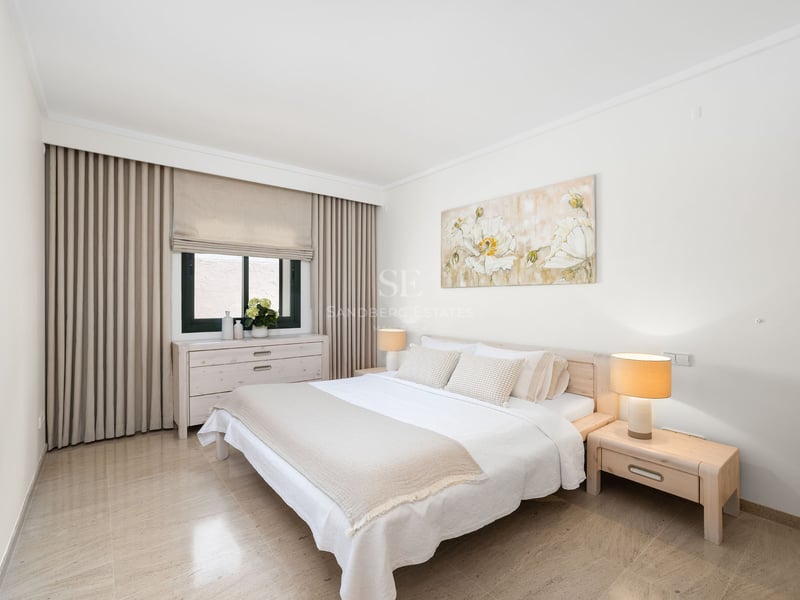 Chambre lumineuse avec lit blanc, tables de chevet en bois, tableau floral et sol en carrelage beige.