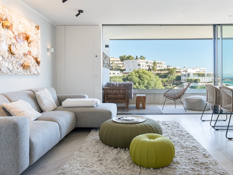 Helles, modernes Wohnzimmer mit grauem Sofa und grünen Sitzpoufs, bodentiefen Fenstern und Blick auf das Meer.